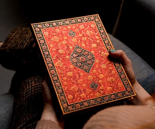 A captivating product image by Tabula Rasa Social Stationers, showcasing a Paperblanks "Layla" journal. The journal's cover features a vibrant red-orange base with intricate gold and black ornate patterns, epitomizing Paperblanks' exquisite artistry and craftsmanship inspired by diverse cultures. Held gently in hands, the image highlights the journal's tactile appeal and premium quality. Designed with durable, eco-friendly materials and high-quality, acid-free paper, it's ideal for writing, sketching, and transforming everyday note-taking into a celebration of creativity and heritage.