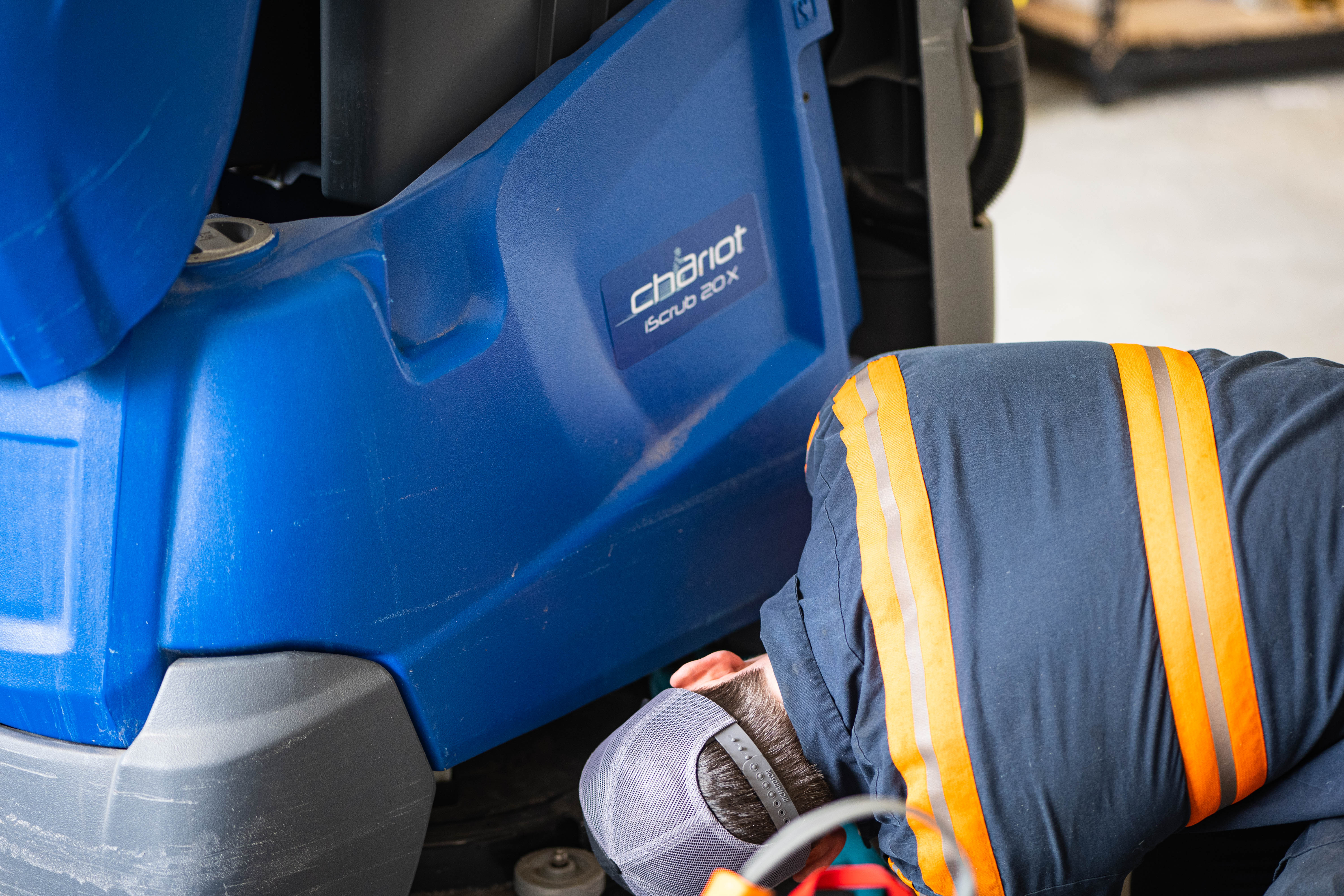 A WaySolutions field technician performs a detailed visual and mechanical inspection on the lower chassis of a blue Kärcher Windsor Chariot iScrub 20X stand-on floor scrubber. By thoroughly evaluating the undercarriage and scrub deck, our Layton-based team catches minor wear and tear before it leads to costly facility downtime for supermarkets and retail centers.