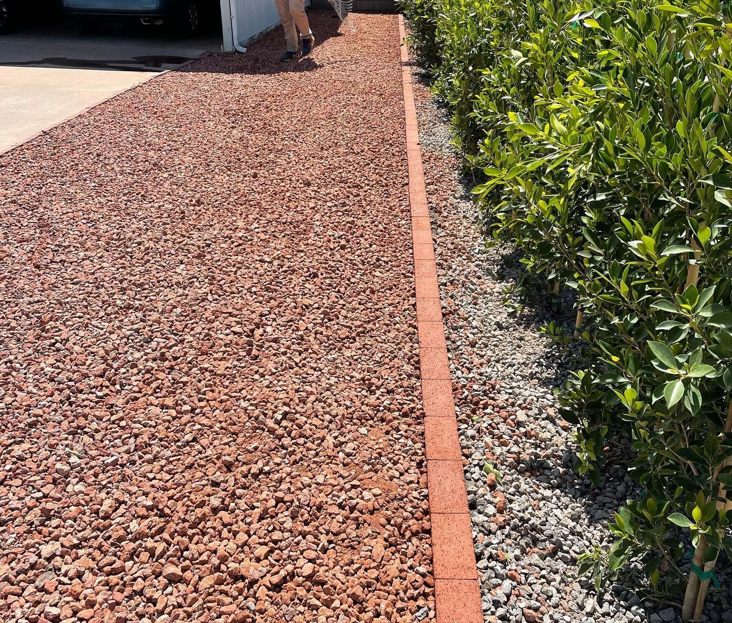 A landscaped side yard with red gravel, a concrete driveway, green shrubs along the edge, and a person walking.