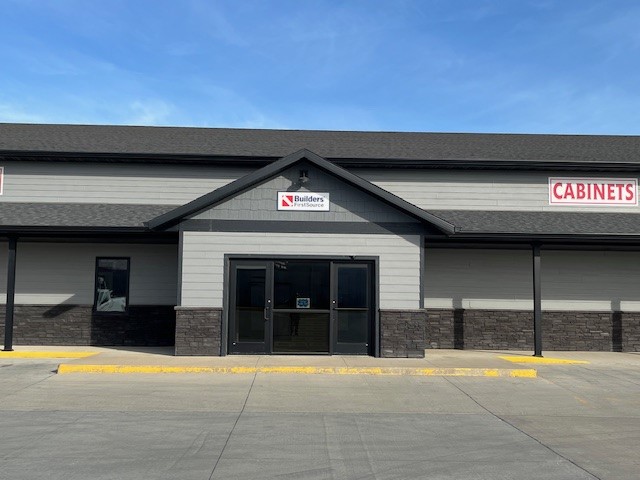 Exterior view of the Builders FirstSource store in Spencer, featuring a modern gray building facade with contrasting dark stone accents along the lower portion. The building has a clean, symmetrical design with large glass double doors at the center and bold red signage for 'Cabinets' on the right. The backdrop highlights a bright blue sky with no clouds, enhancing the fresh and inviting appearance of the store.