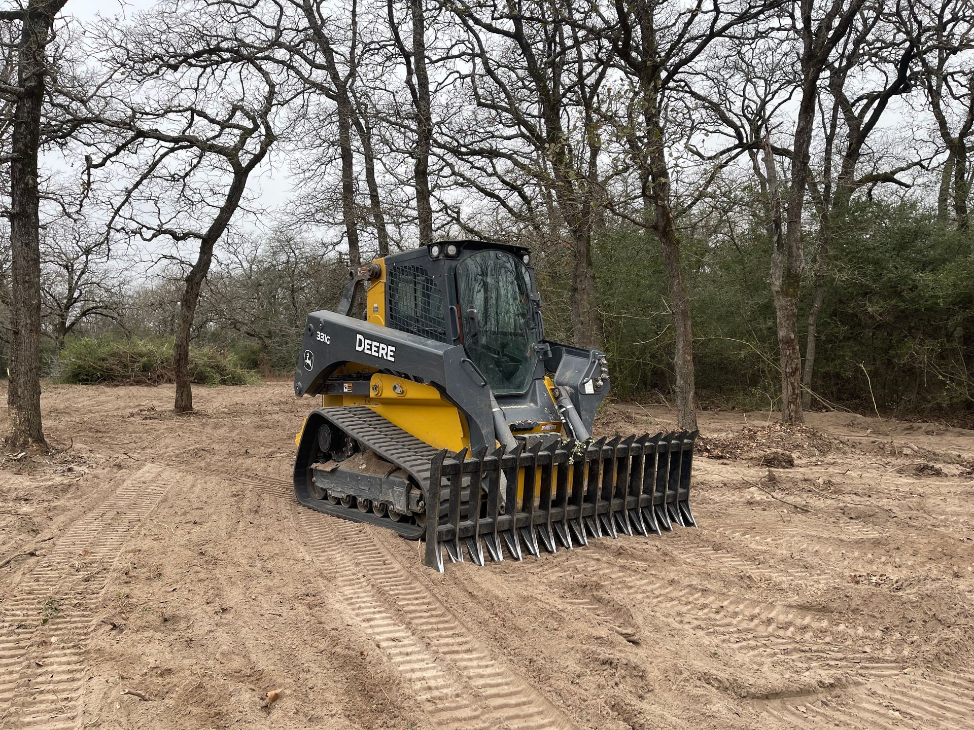 Tracked skid steer equipped with rake attachment smoothing soil on cleared land, highlighting grading, finishing, and land preparation services for landscaping, construction sites, or property development in wooded surroundings.