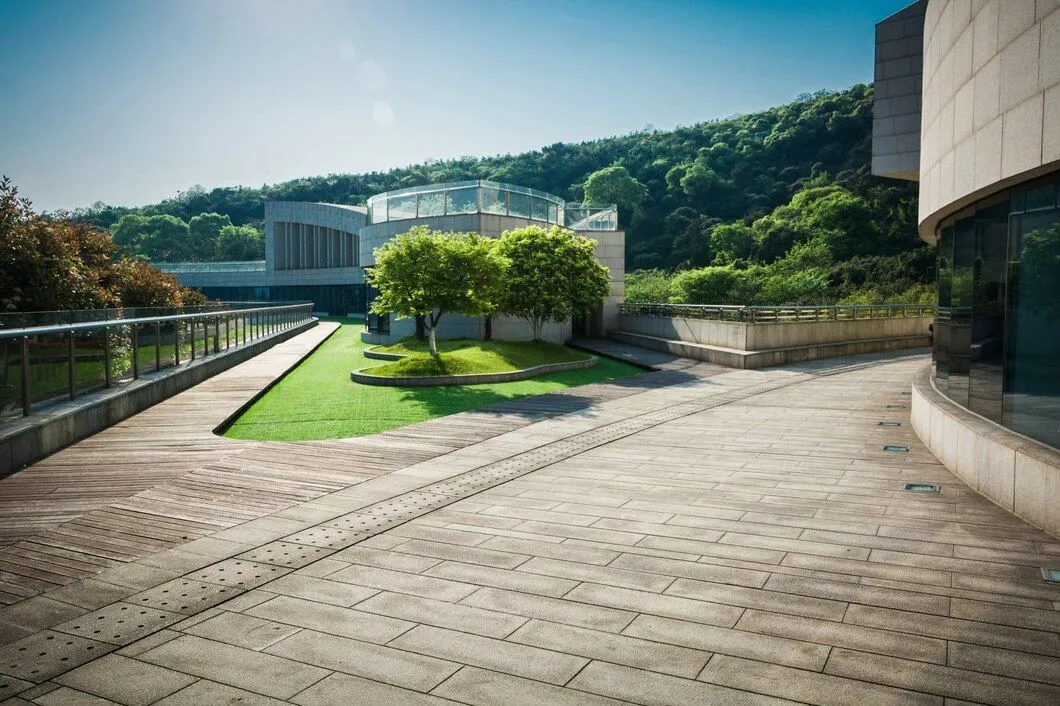 A wide shot of a modern building complex with curved architecture, surrounded by lush greenery and a clear blue sky. The foreground features a wooden pathway and a paved area with tactile paving. In the mid-ground, there's a section of artificial turf, neatly manicured, and several trees. The background shows a dense forest covering a hillside.