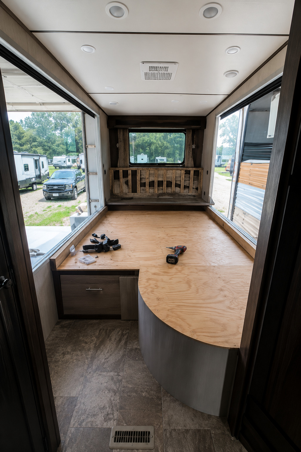 RV interior undergoing renovation with furniture removed, exposing a plywood platform and cabinetry, large open side sections, ceiling lights, ventilation, scattered tools, and surrounding campground vehicles clearly visible outside nearby.