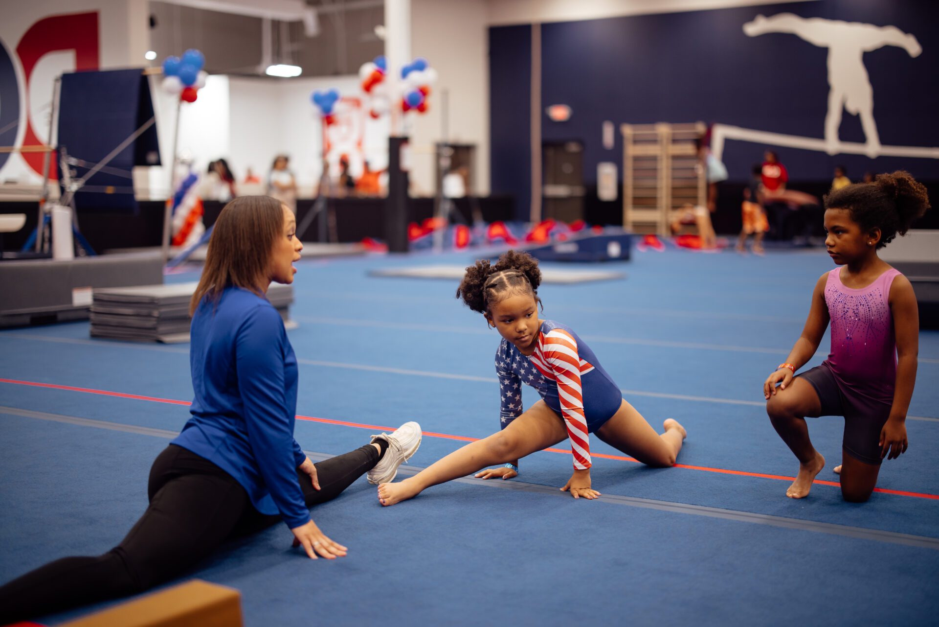 gymnastics coach teaching two young girls how to do a gymnastics move on a blue mat