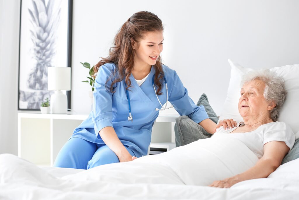 A young nurse in blue scrubs with a stethoscope around her neck is tending to an elderly woman in bed. The nurse is leaning over the bed and gently placing her hand on the woman's shoulder, speaking to her kindly. The room appears to be a bedroom or hospital room, with a white bed, white linens, and a bedside table with a lamp and a plant. A framed picture is visible on the wall in the background.