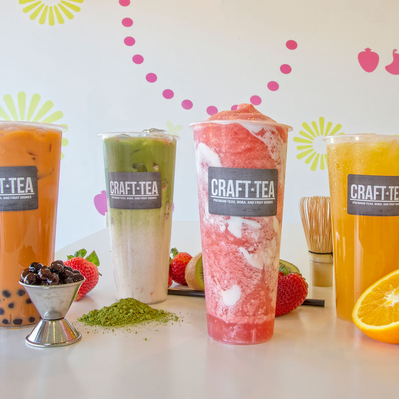 Four colorful drinks are arranged on a white surface, with fresh fruit and ingredients in the foreground. From left to right, the drinks appear to be Thai tea with boba, a green tea beverage, a strawberry smoothie, and an orange-flavored drink. The drinks are in clear plastic cups with "CRAFT-TEA" labels. Strawberries, kiwi slices, a small metal bowl of boba pearls, and matcha powder are scattered around the drinks. A whisk for matcha preparation is visible behind the drinks. The background features a white wall decorated with pink polka dots and yellow sunburst patterns.