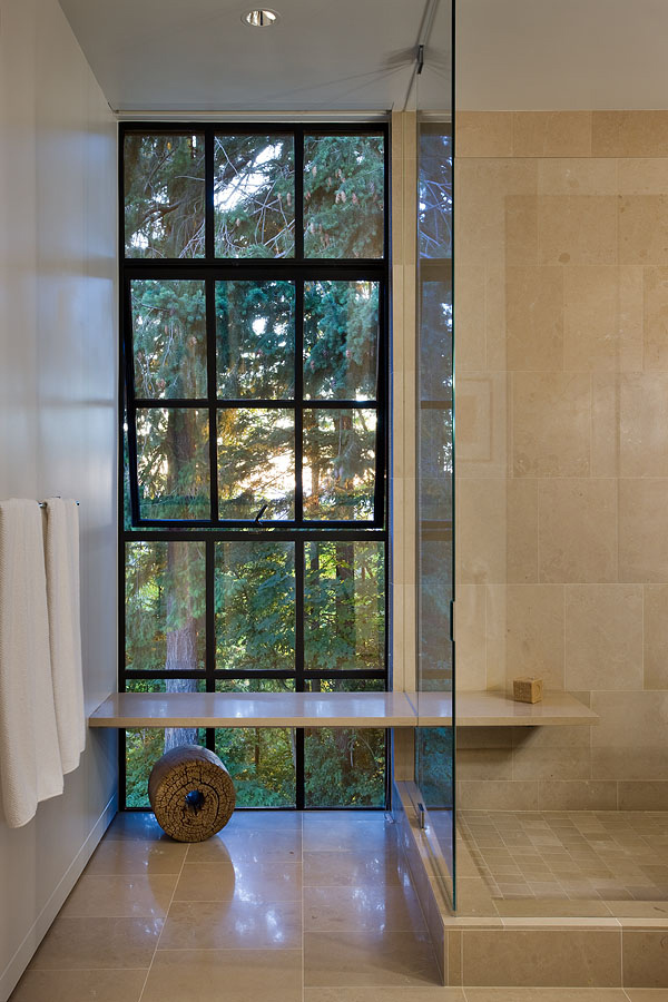 Modern bathroom featuring a large, black-framed grid window beside a tiled shower. The window overlooks trees, while a minimalist bench, light-colored tile, towel, and polished flooring complete the space.