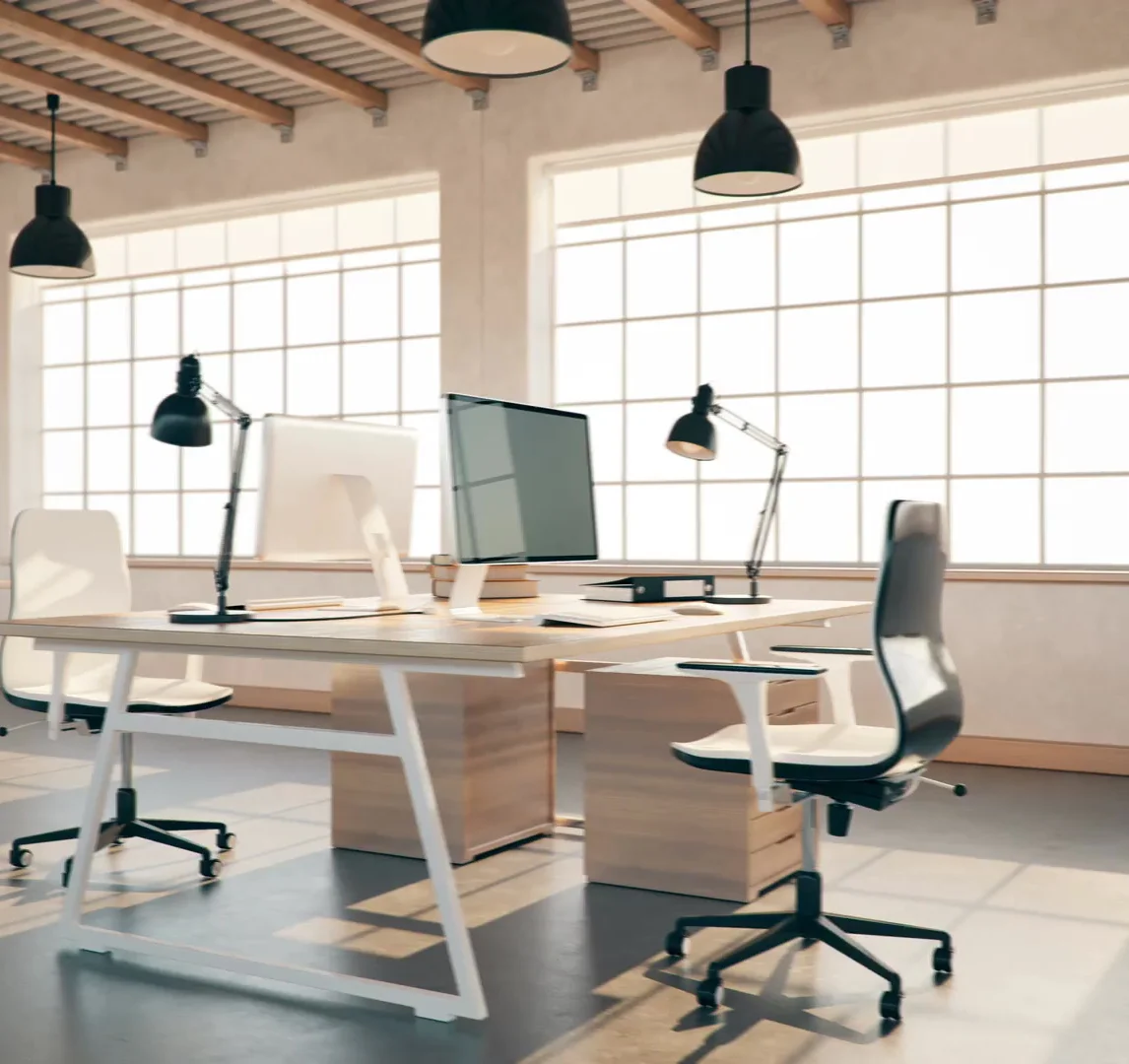 A modern office space with two desks, computers, and chairs. The desks are light wood with white metal legs. Each desk has a computer monitor, a desk lamp, and office supplies. The space is filled with natural light from large, grid-patterned windows.