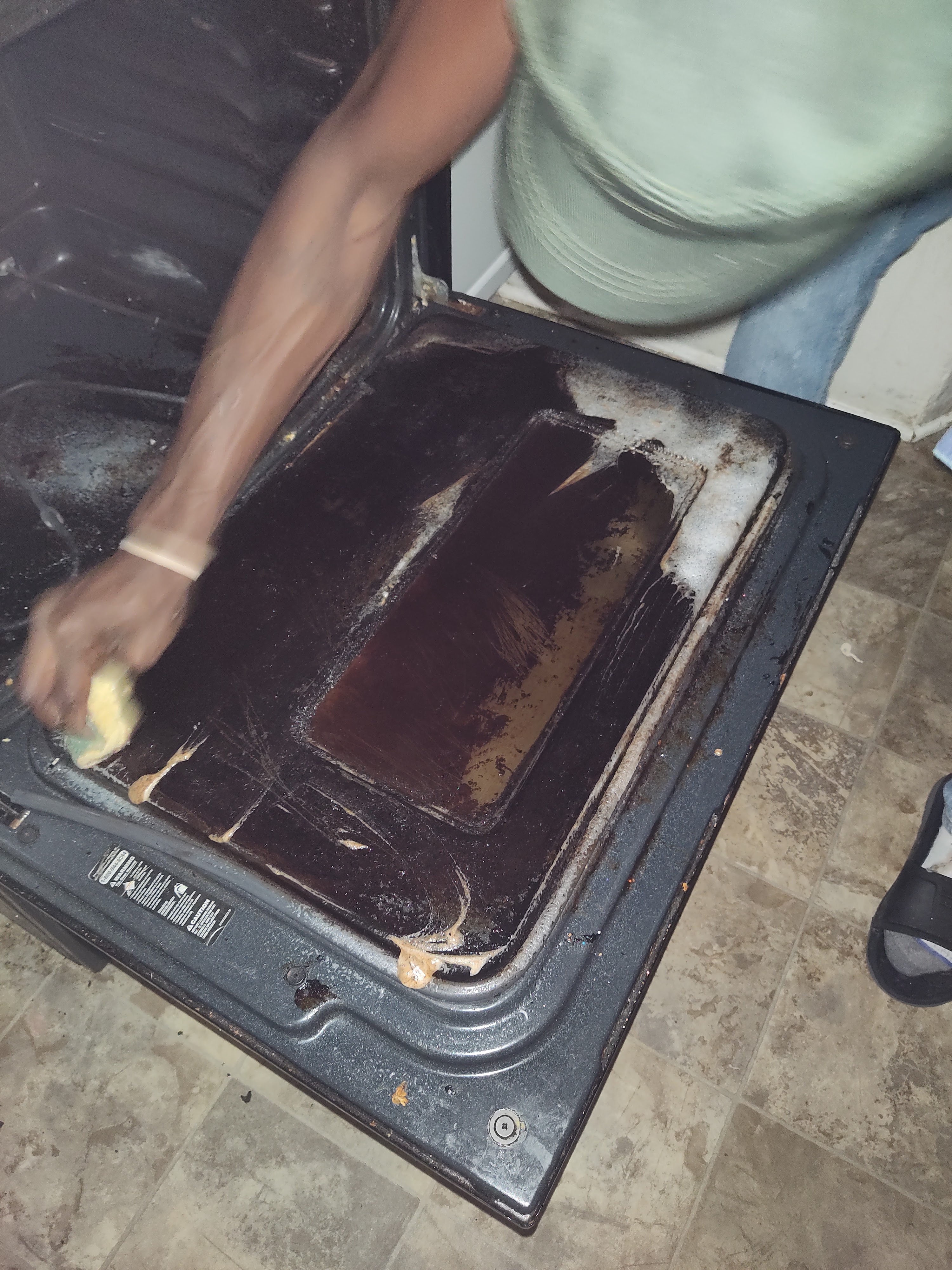 A person's arm is visible cleaning the inside of a dark-colored oven with a yellow sponge. The oven interior appears grimy with dark residue and some lighter-colored cleaning product or residue. The person is wearing a light green shirt and blue jeans, and a light-colored wristband. The oven is on a tiled floor with a pattern of light browns and grays. A black sandal is visible on the right side of the frame.