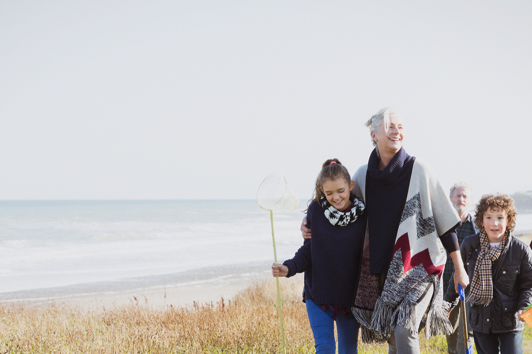 A joyful family strolls along a grassy beach path, with the ocean in the background. A young girl holds a fishing net, smiling alongside an older woman.