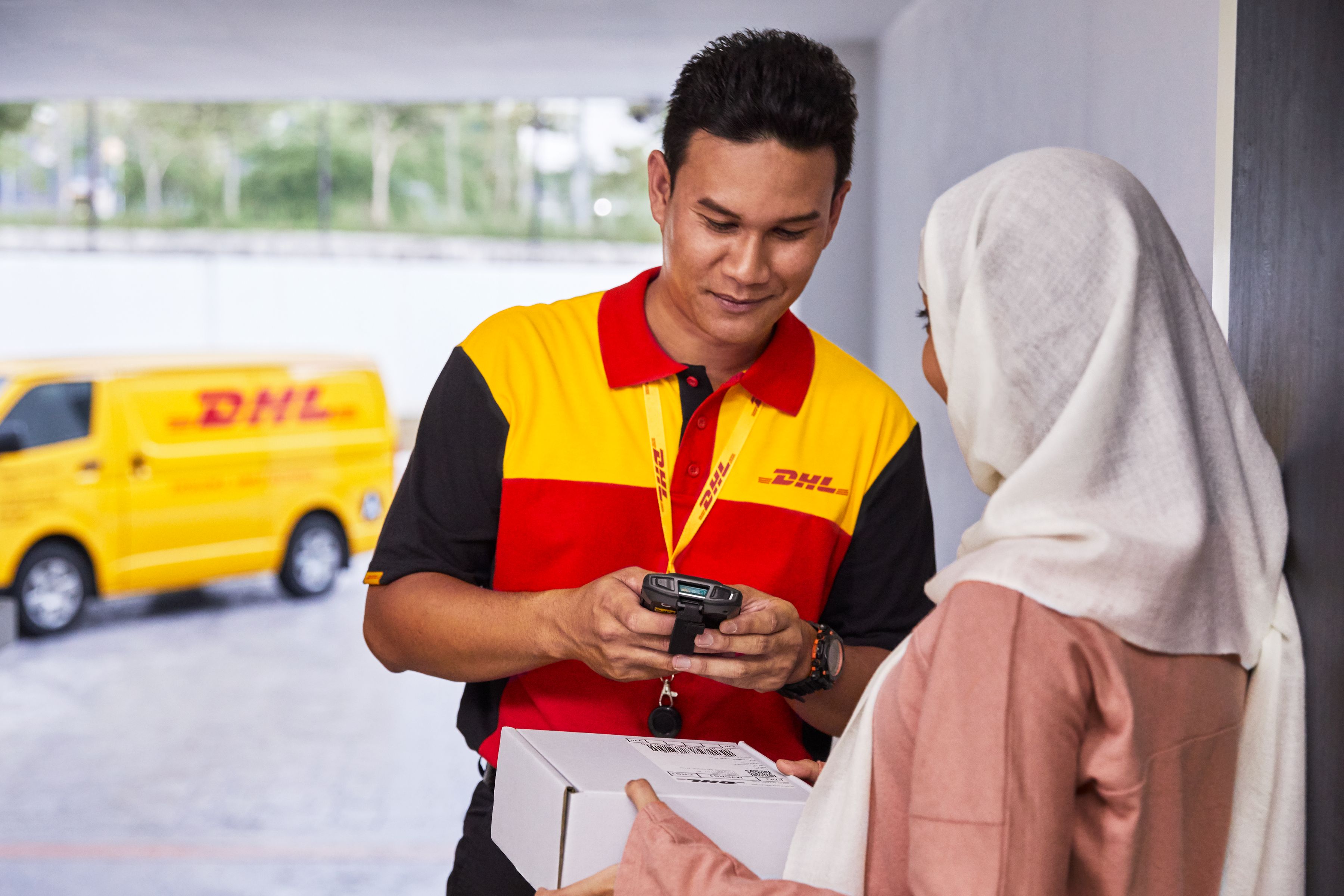 A DHL Express courier in a red and yellow uniform is handing over a parcel to a customer at a doorway while using a handheld device for delivery confirmation. A yellow DHL van is visible in the background.
