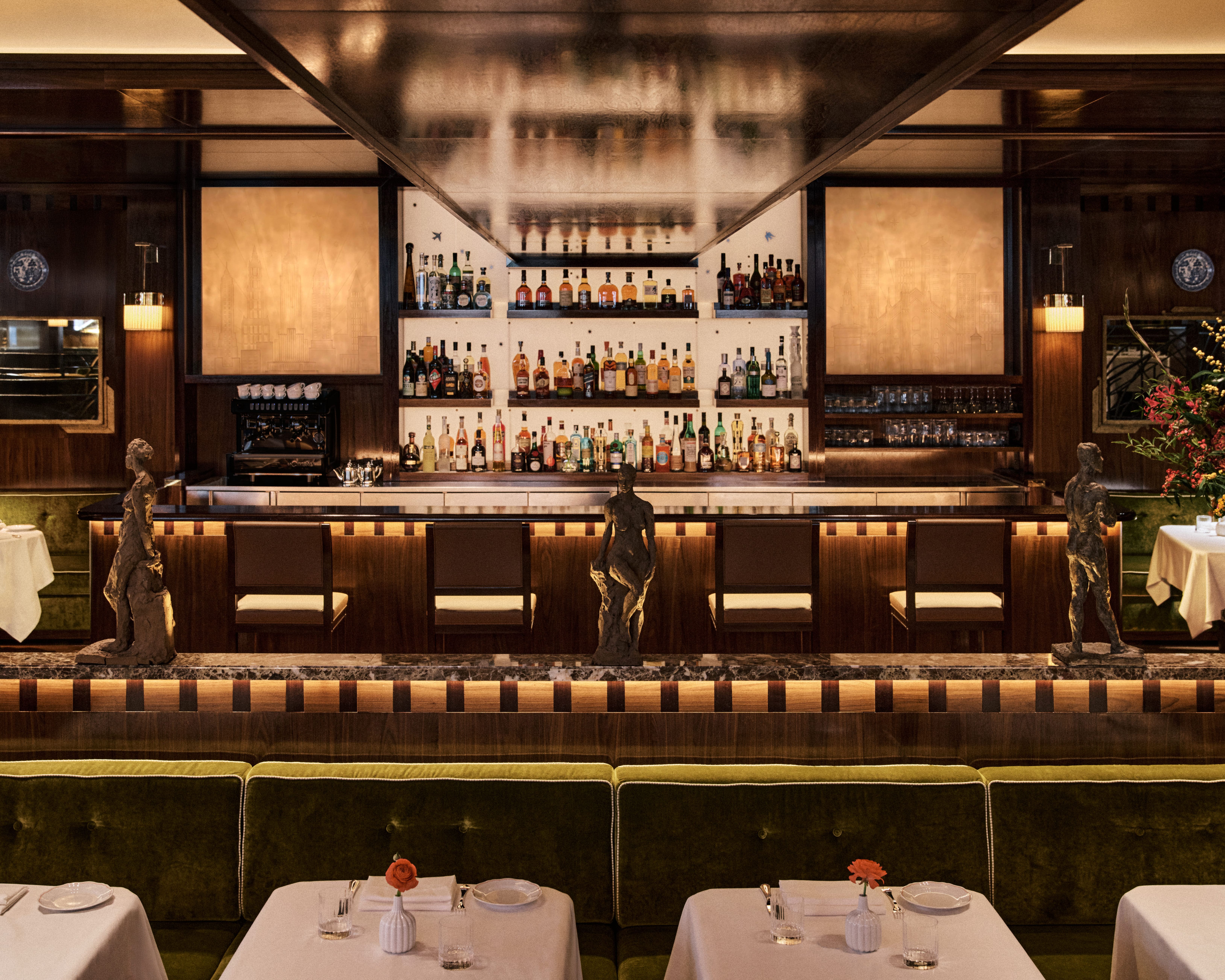 Sophisticated bar interior with a long, backlit counter lined with bar stools, shelves of spirits beneath a mirrored canopy, sculptural figures along the bar front, and plush green banquette seating set with white tablecloths and single flowers.