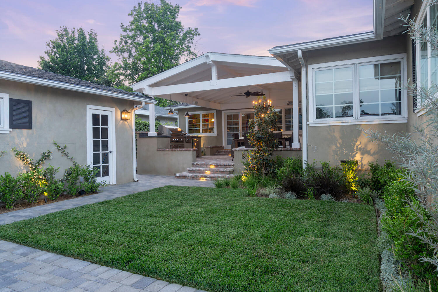 Serene stucco home courtyard at dusk, with lush lawn, flowering gardens, covered patio, warm lights, and inviting steps to the entry.