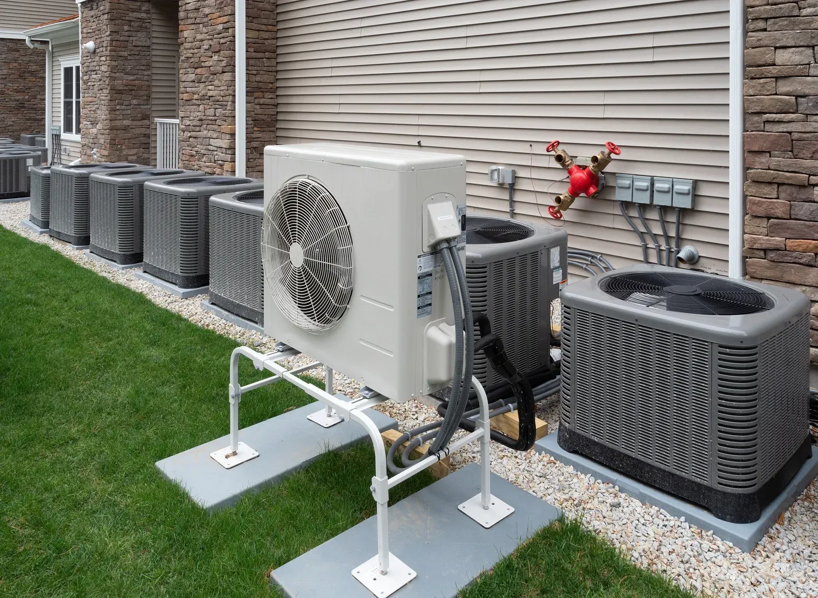 A row of grey AC condensers lines a building with beige siding and stone columns. In the foreground, a slim white unit sits elevated on a metal stand above concrete pads.