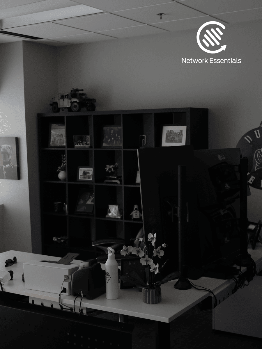 A black and white photograph of an office desk setup, featuring dual monitors, a printer, a water bottle, and a small potted orchid. Behind the desk, a dark bookshelf is filled with framed photos and decorative items, including a model vehicle on the top shelf. The Network Essentials logo is visible in the upper right corner.