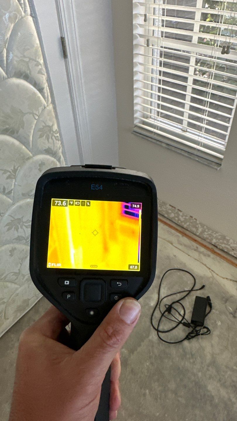 A hand holds a FLIR E54 thermal imaging camera, displaying a heat map of a room. To the left, a mattress leans against a wall and a door frame. A window with white blinds is visible on the right, and a power adapter with a coiled cord lies on the floor.