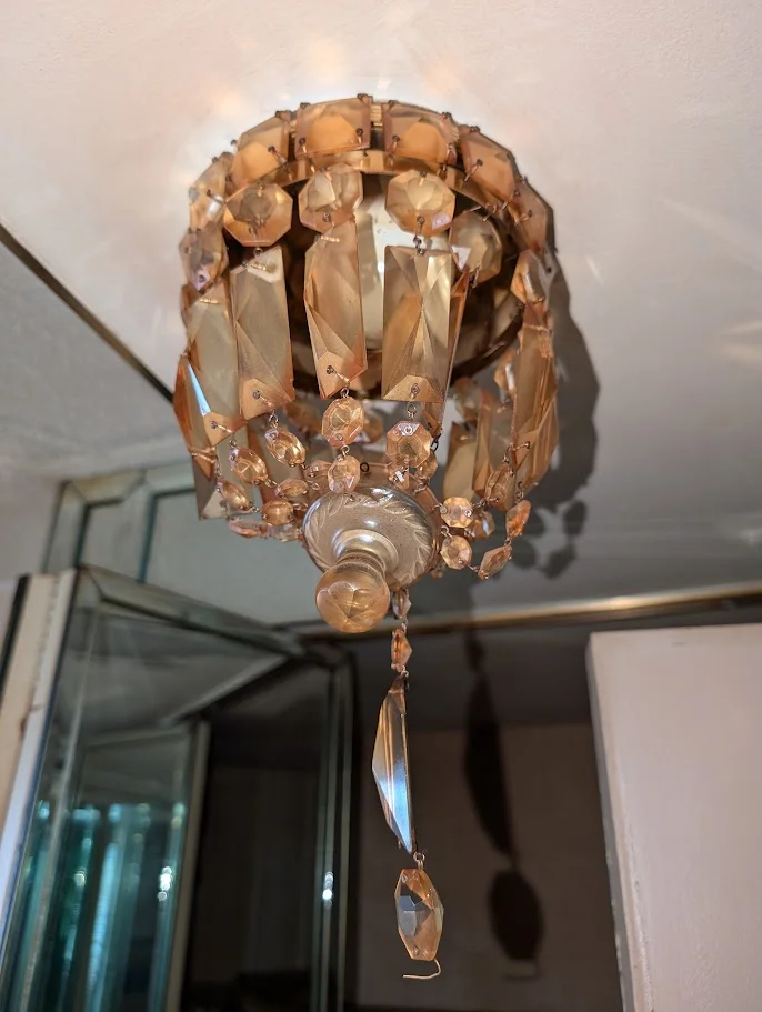 A close-up, low-angle shot of a decorative chandelier with many amber-colored crystals hanging from its metal frame. The chandelier appears to be illuminated by a light source within it, casting shadows on the ceiling and the wall behind it.