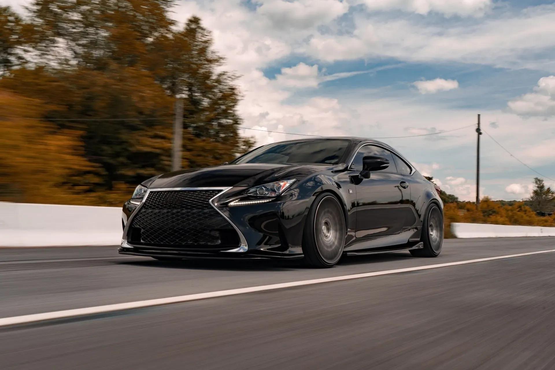 A black Lexus RC coupe drives down a paved road, with trees and a cloudy sky in the background. The car is captured in motion, with the wheels blurred.