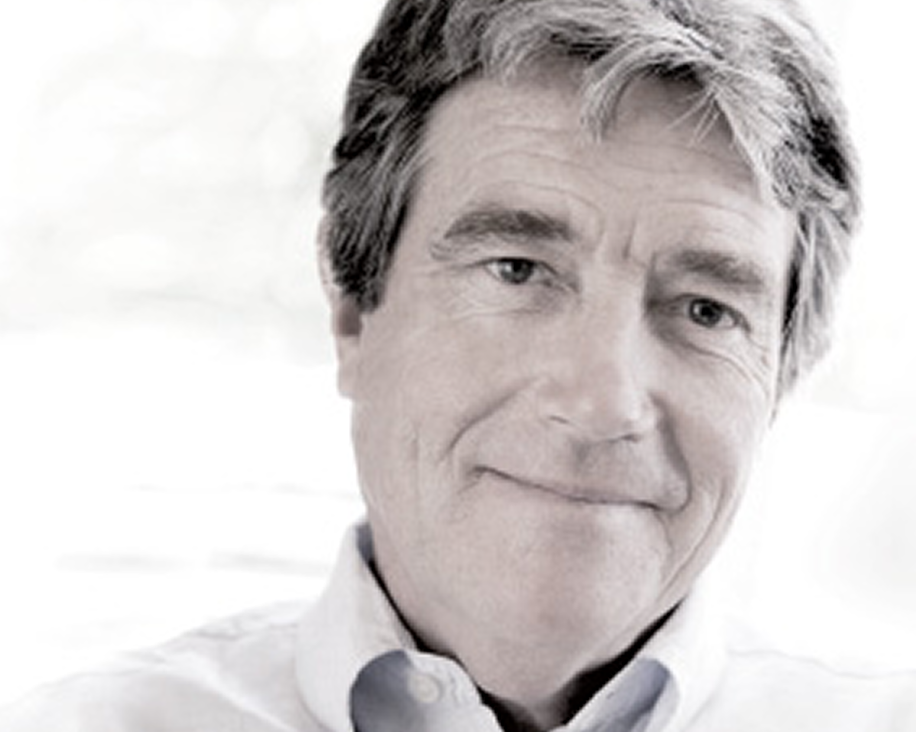 A person with light-colored hair wearing a white shirt, sitting indoors with a bright, blurred background.