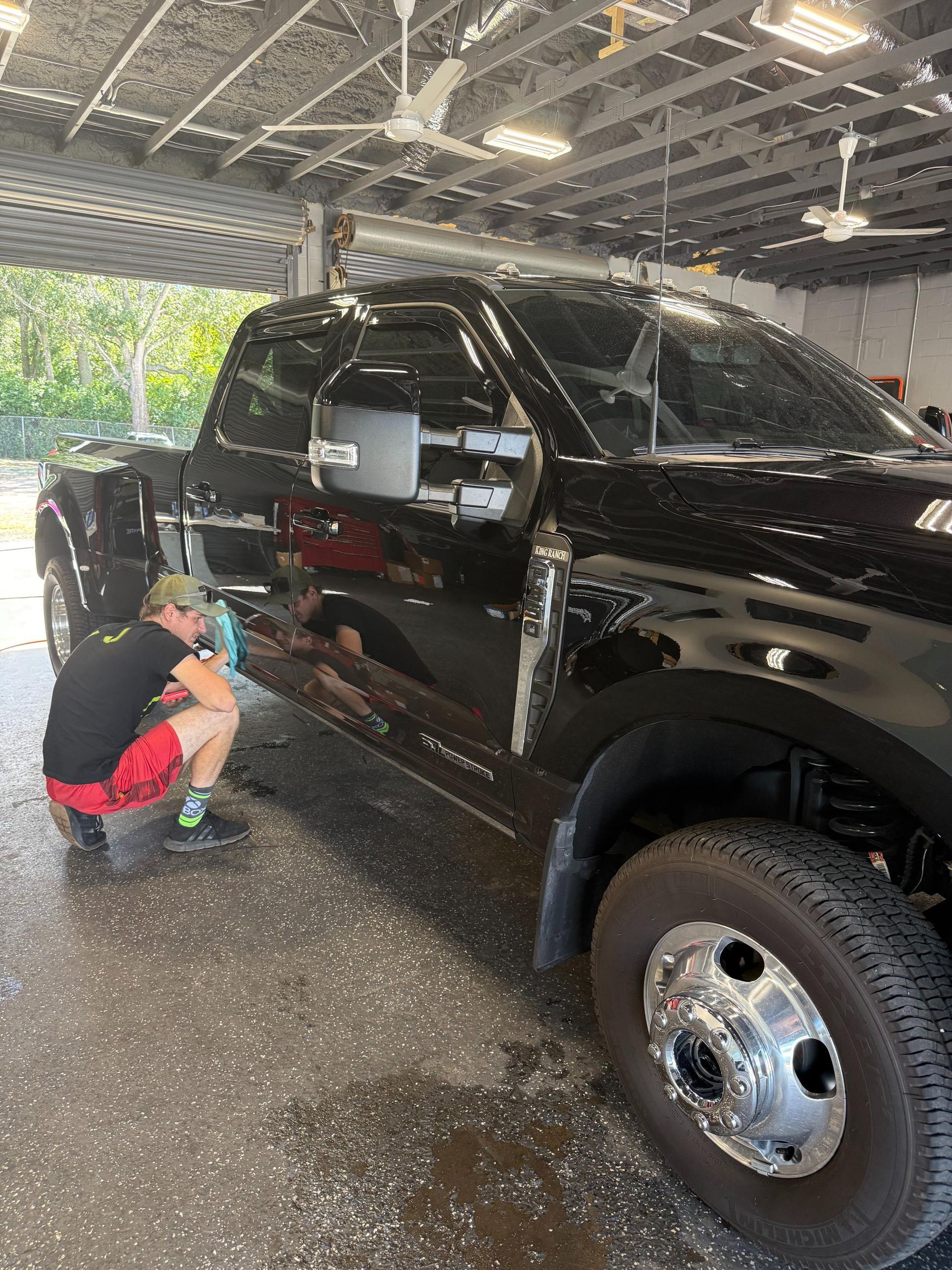 A technician kneels down to clean and detail the lower side panel of a large, black, dual-rear-wheel pickup truck, suggesting professional detailing or paint correction service is being performed in a sunny garage bay.