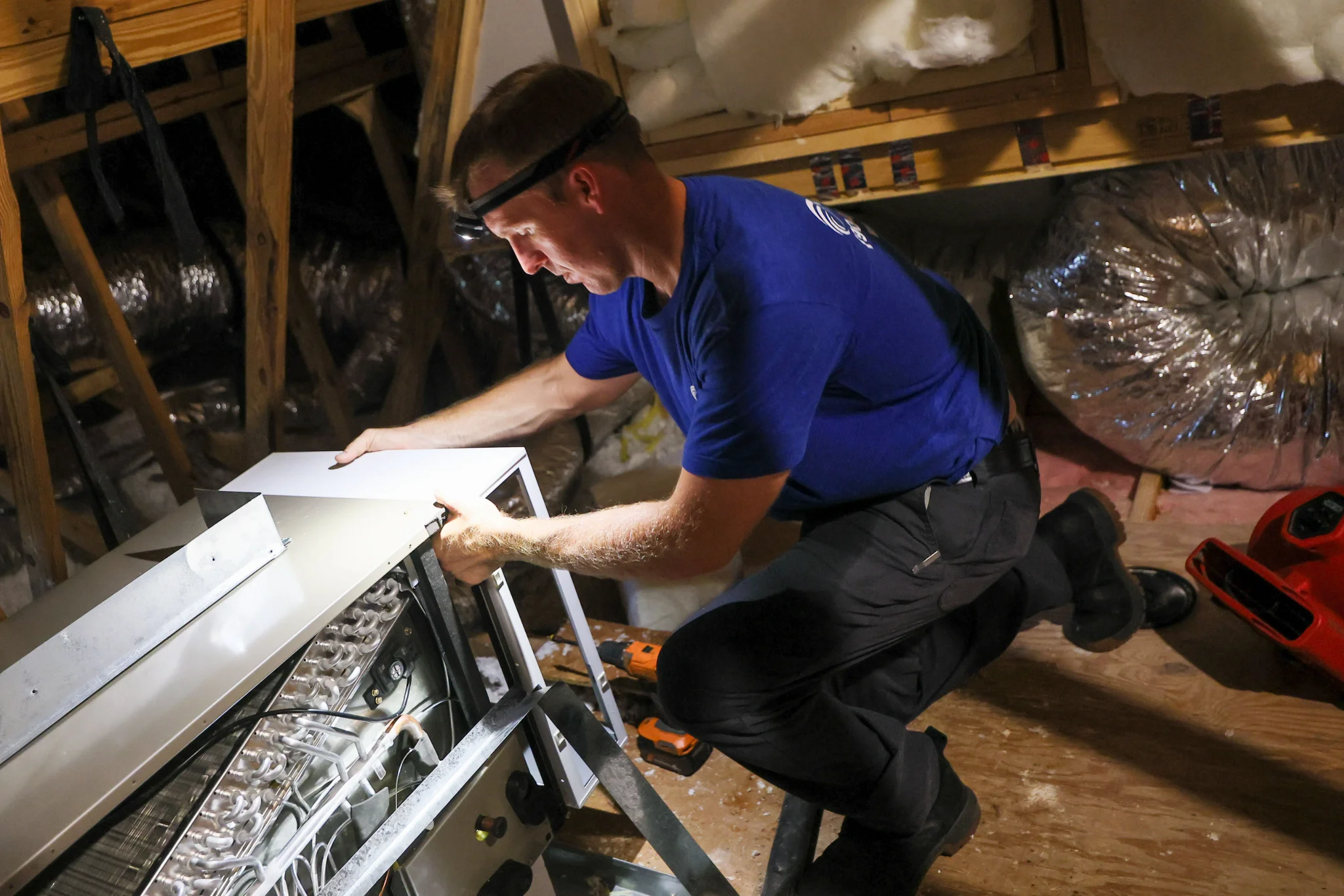 A man in a blue shirt and headlamp is working on an HVAC unit in an attic. He is kneeling and holdin 