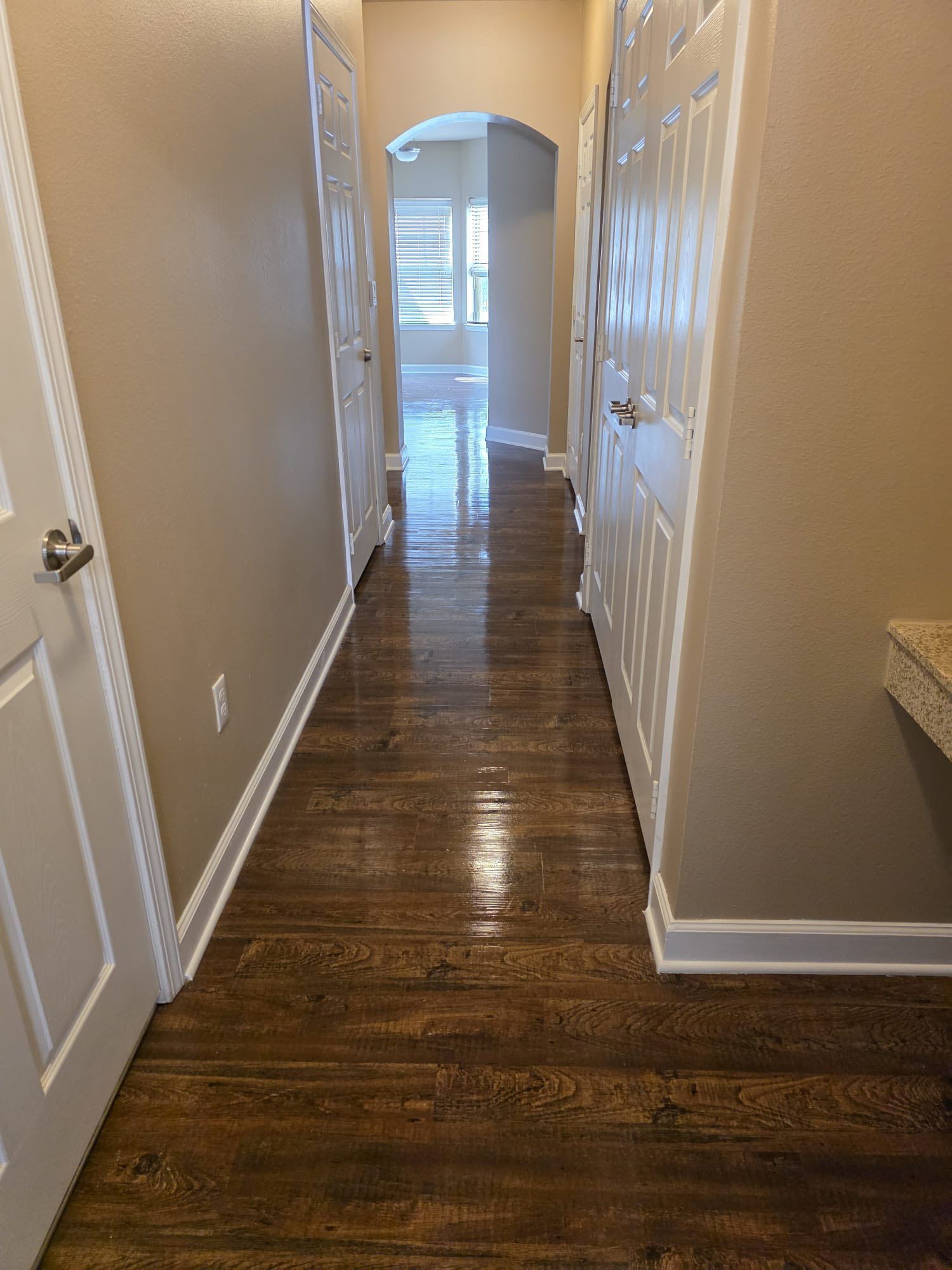 A residential hallway with dark wood flooring, beige walls, and white doors leading to different rooms. An arched opening at the end reveals a bright space with windows and natural light.