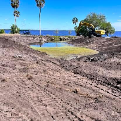 An excavator shaping the dark dirt banks of a large pond excavation site near a lake with palm trees.