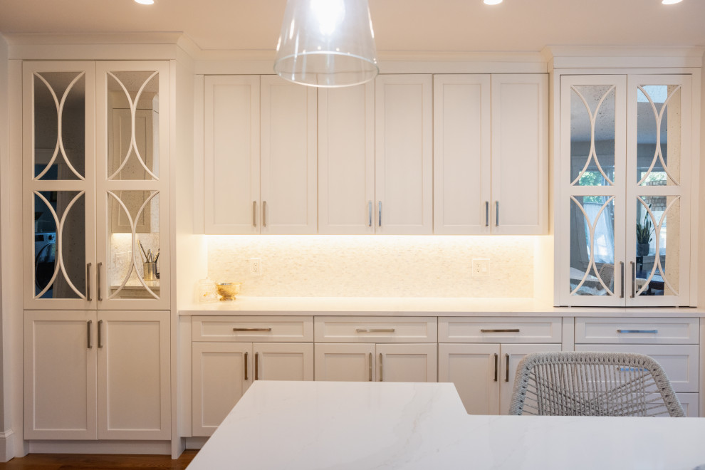 A modern kitchen with white cabinetry, a white marble countertop, and subtle under-cabinet lighting. The cabinets feature decorative mirrored glass panels with an art deco-inspired design. A woven chair is visible in the foreground, suggesting a seating area within the kitchen. The overall aesthetic is clean, bright, and sophisticated.