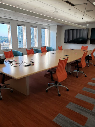 A bright and spacious conference room featuring a long, light-colored wooden table surrounded by modern orange office chairs with ergonomic designs. Large windows line one side of the room, offering a panoramic view of a cityscape and filling the space with natural light. The room also includes a wall-mounted flat-screen television for presentations, and the warm-toned hardwood flooring adds a welcoming touch.