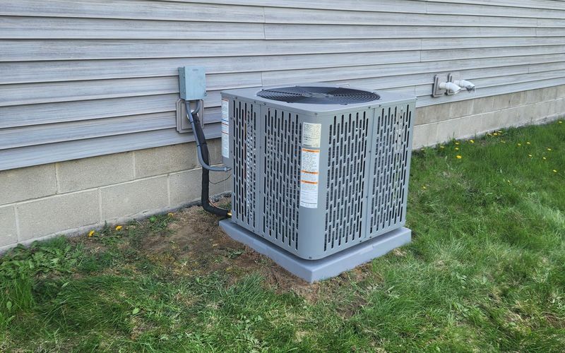 A close-up shot of a modern, grey residential air conditioning condenser unit installed on a level pad. The unit features a perforated protective grille and is connected to a home&rsquo;s exterior.