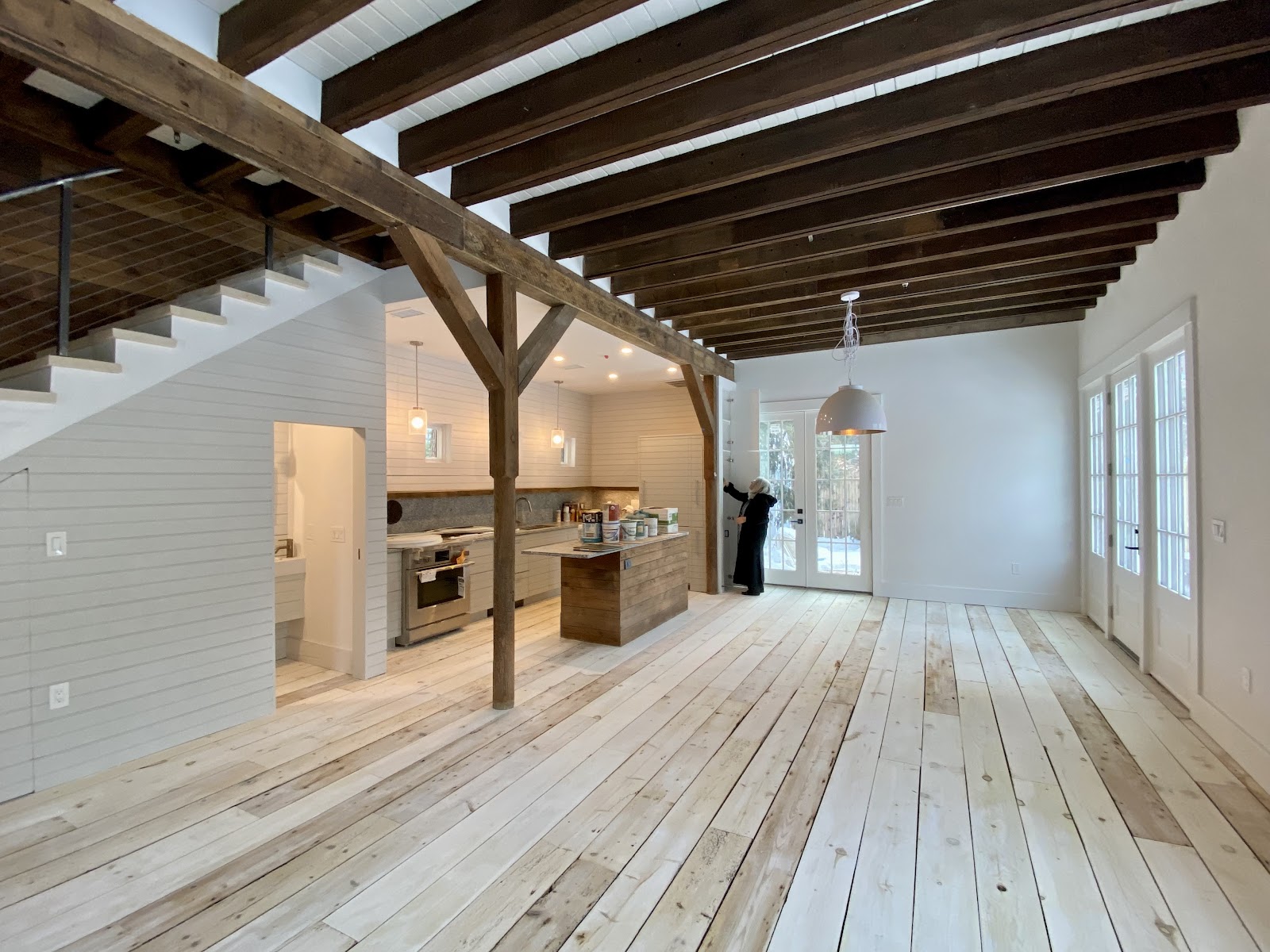 A person in a black coat is standing by a door in a room with wooden beams on the ceiling and a wooden floor. There is a kitchen area with cabinets and a stove in the background. Several cans of paint are on a wooden island in the center of the room. The room appears to be under renovation or recently completed.