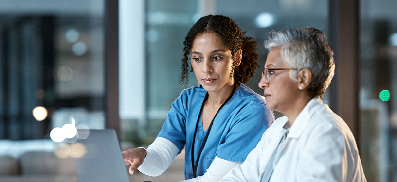 A woman in scrubs and a woman in a lab coat examining data on a screen.