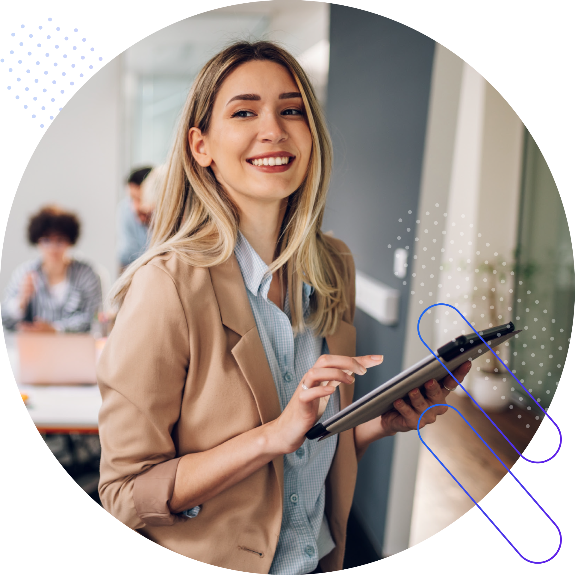 A blonde woman wearing a beige blazer and a blue plaid shirt is smiling and looking to her left while holding a tablet in an office setting. In the background, two other people are blurred out, one is looking at a laptop.