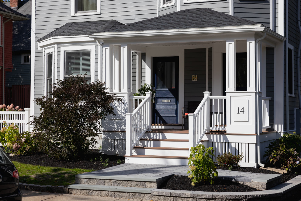 A charming front porch with a dark blue door and white railings leads up to a grey clapboard house. The number "14" is displayed prominently on a white panel next to the stairs. Landscaping includes dark mulch, a dense dark-leaved bush, and flowering hydrangeas. A portion of a black car is visible in the lower left corner.