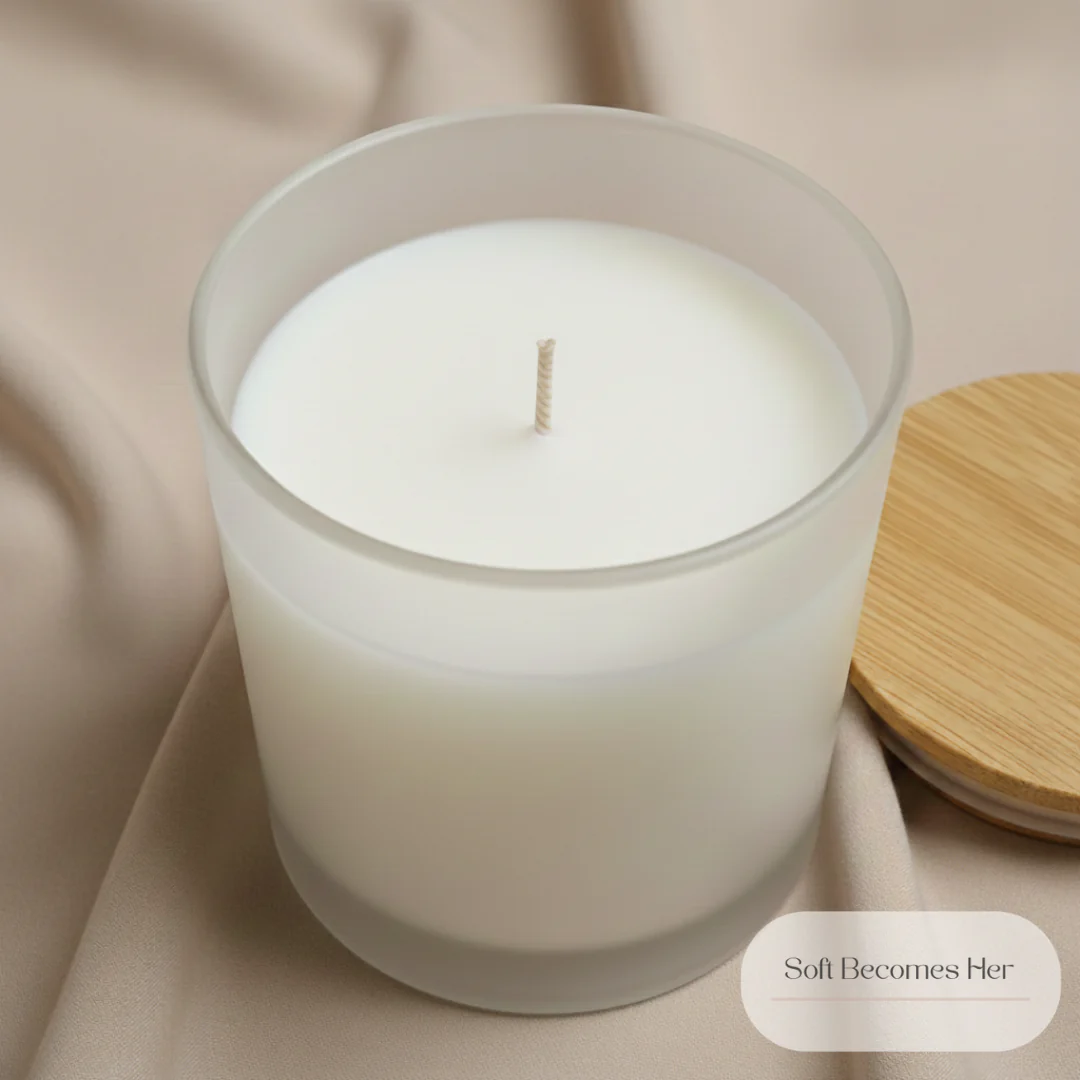 A close-up, overhead shot features a white candle in a frosted glass container. The candle is unlit, with a single wick positioned centrally. The container rests on a soft, beige fabric that is draped and folded. To the right of the candle, a circular wooden lid, possibly bamboo, is partially visible, suggesting a lid for the candle container. A small, oval-shaped graphic element in the bottom right corner contains the text "Soft Becomes Her" in a light brown, elegant font. The overall aesthetic is minimalist and serene, emphasizing the texture and soft colors of the elements.