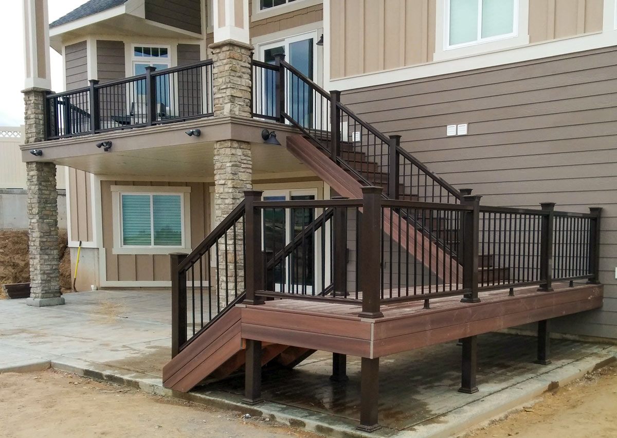 Elevate your home's outdoor living space with this custom multi-level deck project by DeckPro LLC. Featuring durable composite decking in a warm tone, the design connects an upper-level balcony to a lower-level deck and patio via a grand staircase. Dark metal railings and beautiful stone veneer columns add a touch of elegance and structural integrity to this impressive backyard upgrade.