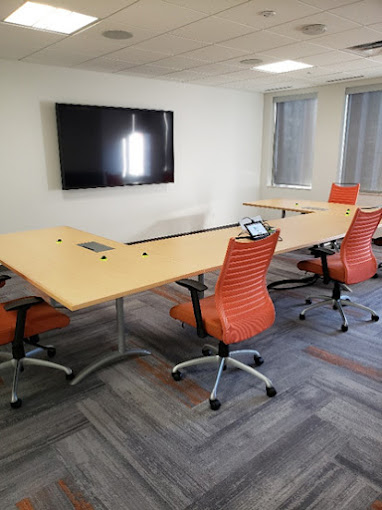 A small, modern meeting room featuring a unique, modular conference table with light wood surfaces and built-in connectivity ports. The table is surrounded by bright orange ergonomic chairs on wheels, providing both comfort and flexibility. The room includes a wall-mounted flat-screen television for presentations or video conferencing, with large windows on one side allowing natural light to enter.