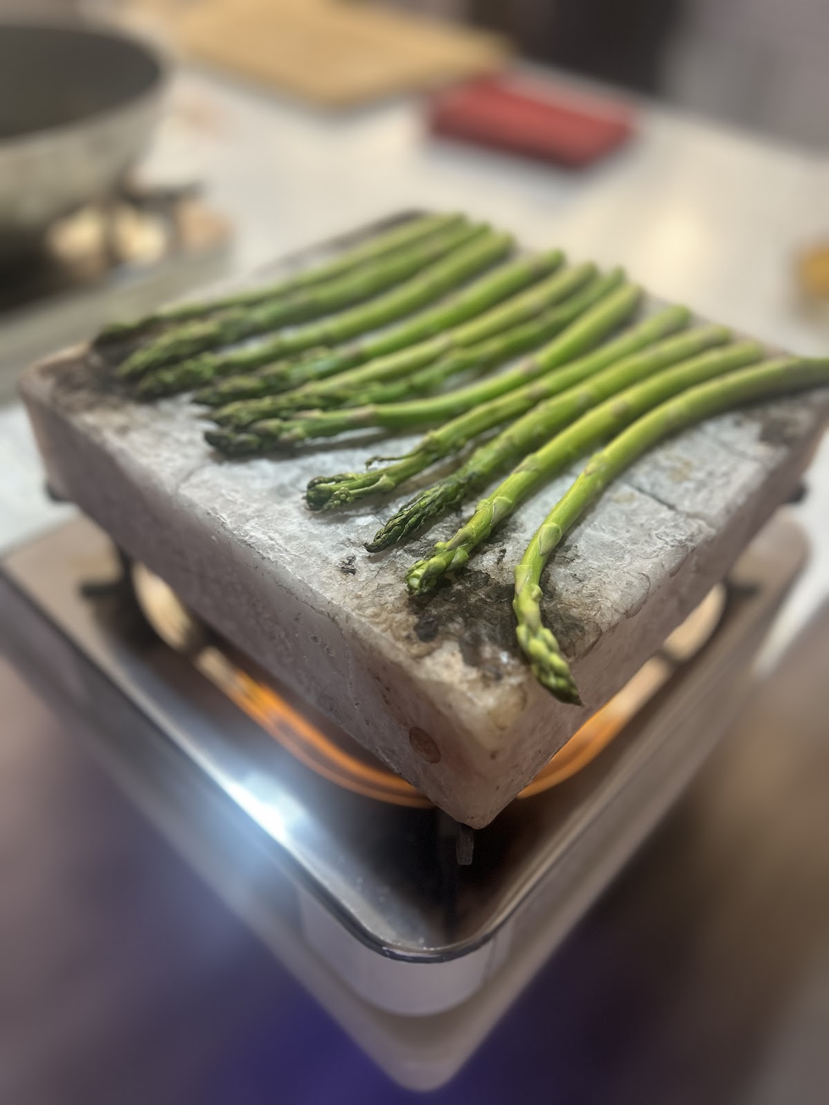 A close-up shot shows a block of Himalayan pink salt with asparagus spears arranged on top, resting on a portable induction cooktop with a blue flame visible.