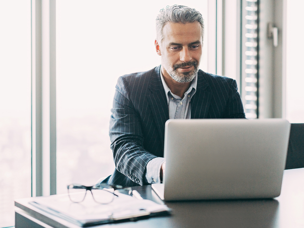 Businessman in a suit working on a laptop at an office desk with eyeglasses and documents. He looks focused, with city skyline visible outside.