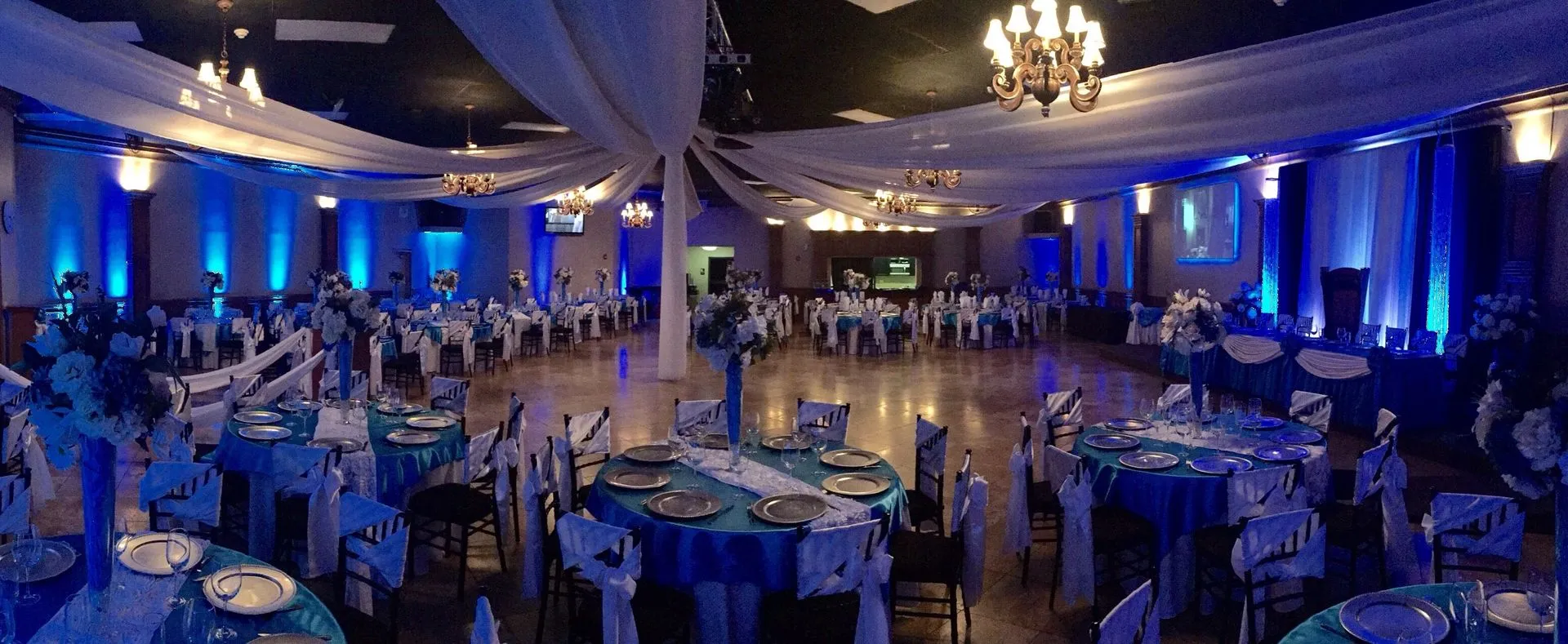 Large reception hall featuring blue uplighting, ceiling drapery, elegant banquet tables, floral cent 