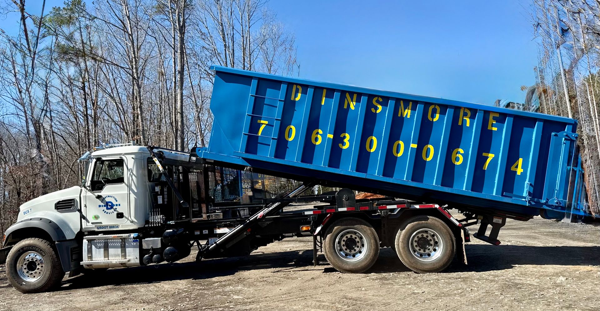 A white roll off truck lifts a large blue dumpster labeled Dinsmore with a phone number. The truck operates on a dirt surface surrounded by trees at a construction site.