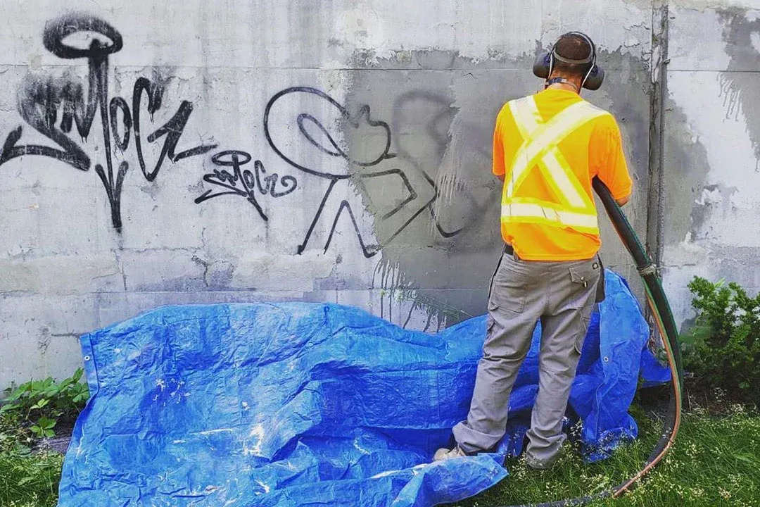Worker removes graffiti from concrete wall using blasting equipment, restoring surface appearance by eliminating paint markings, ensuring clean finish while maintaining structural integrity and preparing area for repainting.