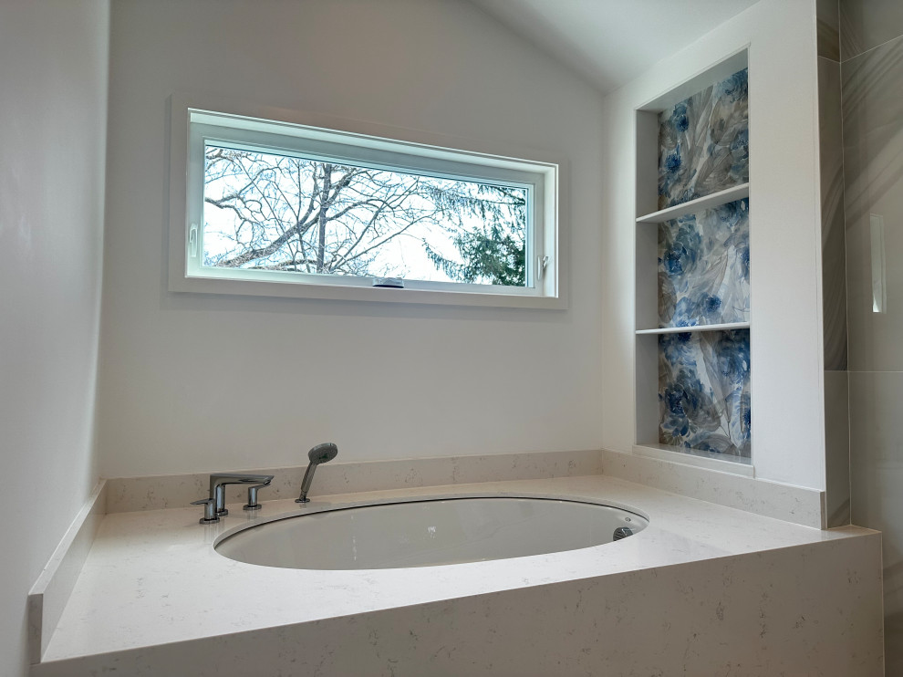 A modern bathroom with a large soaking tub and a picturesque window view of trees. The tub is set into a white countertop, and a sleek chrome faucet and handheld showerhead are visible. To the right, a built-in shelf unit is decorated with floral wallpaper in shades of blue and gray. The overall aesthetic is clean and sophisticated, with a focus on natural light and calming colors.