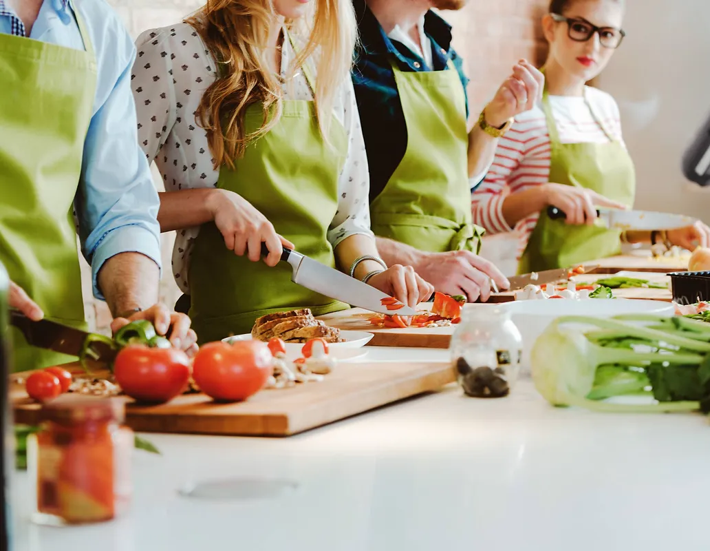A group of four people, two men and two women, are actively engaged in chopping vegetables and preparing food in a kitchen setting. They are all wearing green aprons and appear to be participating in a cooking class or a shared meal preparation. The foreground shows various ingredients like tomatoes, bell peppers, mushrooms, and leafy greens, along with cutting boards and knives. The people are focused on their tasks, with one woman wearing glasses looking towards the camera.