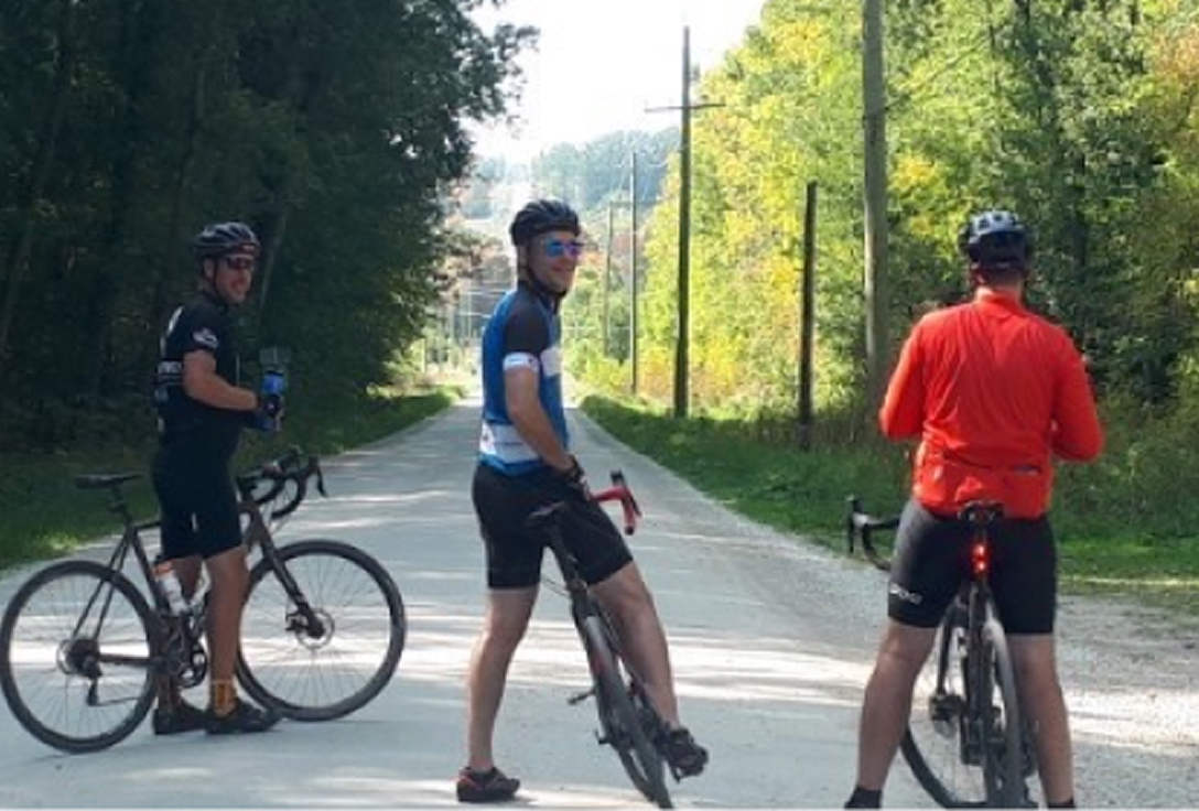 Three cyclists in helmets rest on a sunlit rural road, surrounded by lush trees. The scene conveys a serene and active outdoor vibe.