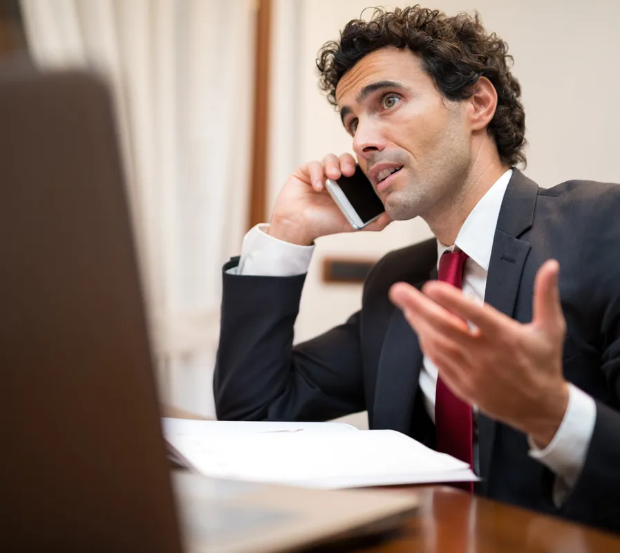 A man in a suit is talking on a cell phone while gesturing with his hand. He is sitting at a desk with a laptop in front of him. The man has curly dark hair and is looking to the right. He is wearing a black suit jacket, a white dress shirt, and a red tie. A laptop is partially visible in the foreground, and papers are on the desk.