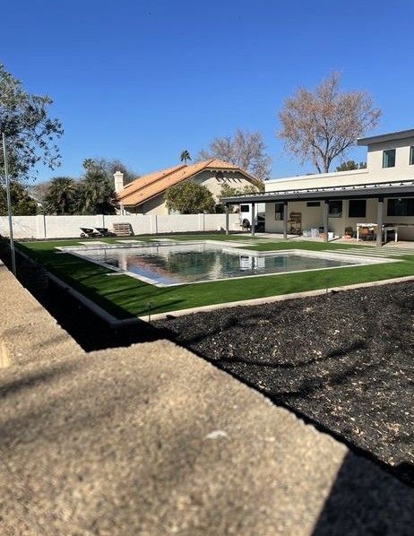 A backyard features a covered pool with artificial grass, modern patio furniture, and neighboring houses under a clear blue sky.