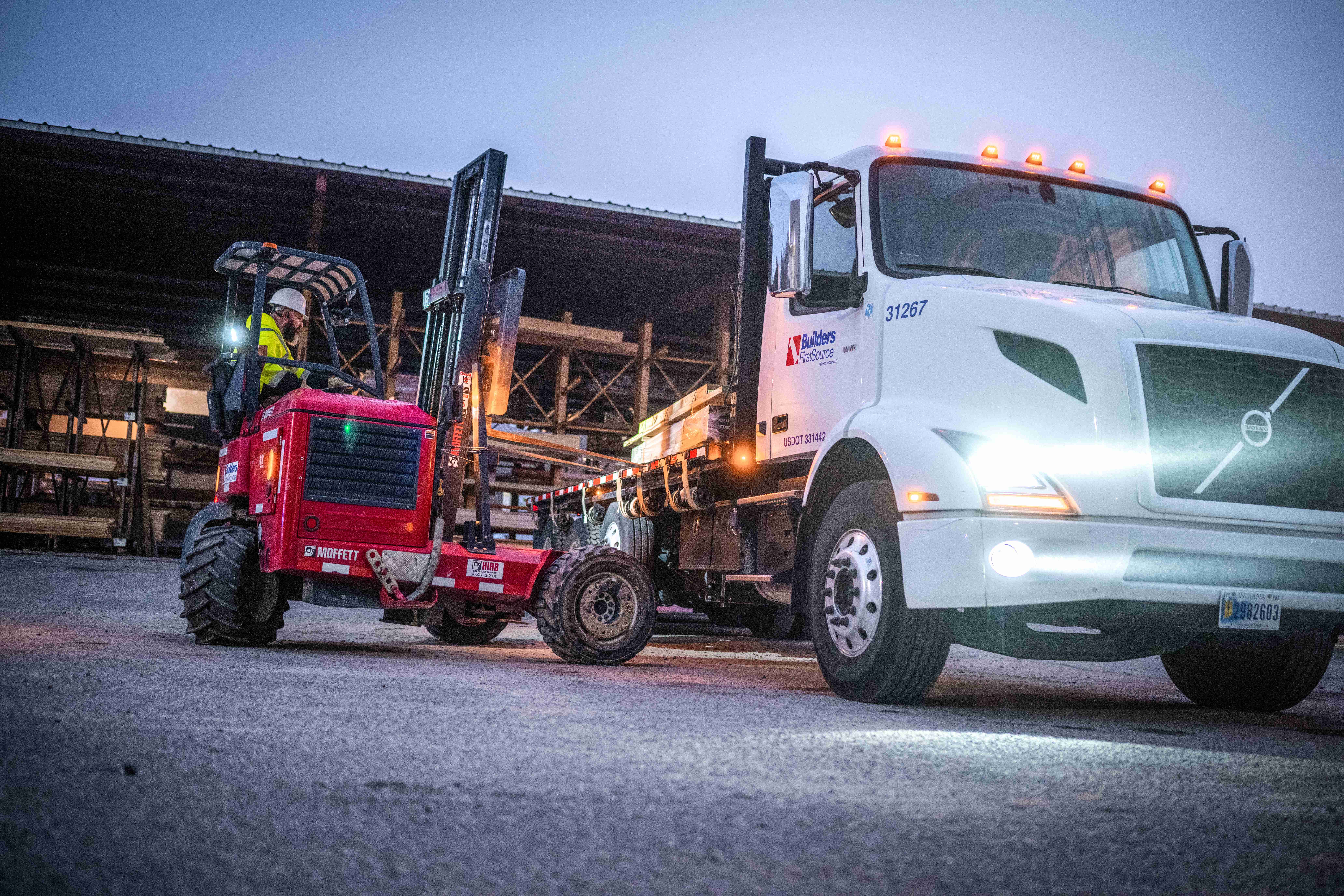 A forklift operator loads lumber onto a Builders FirstSource flatbed truck in a lumber yard, highlighting delivery and material handling services.