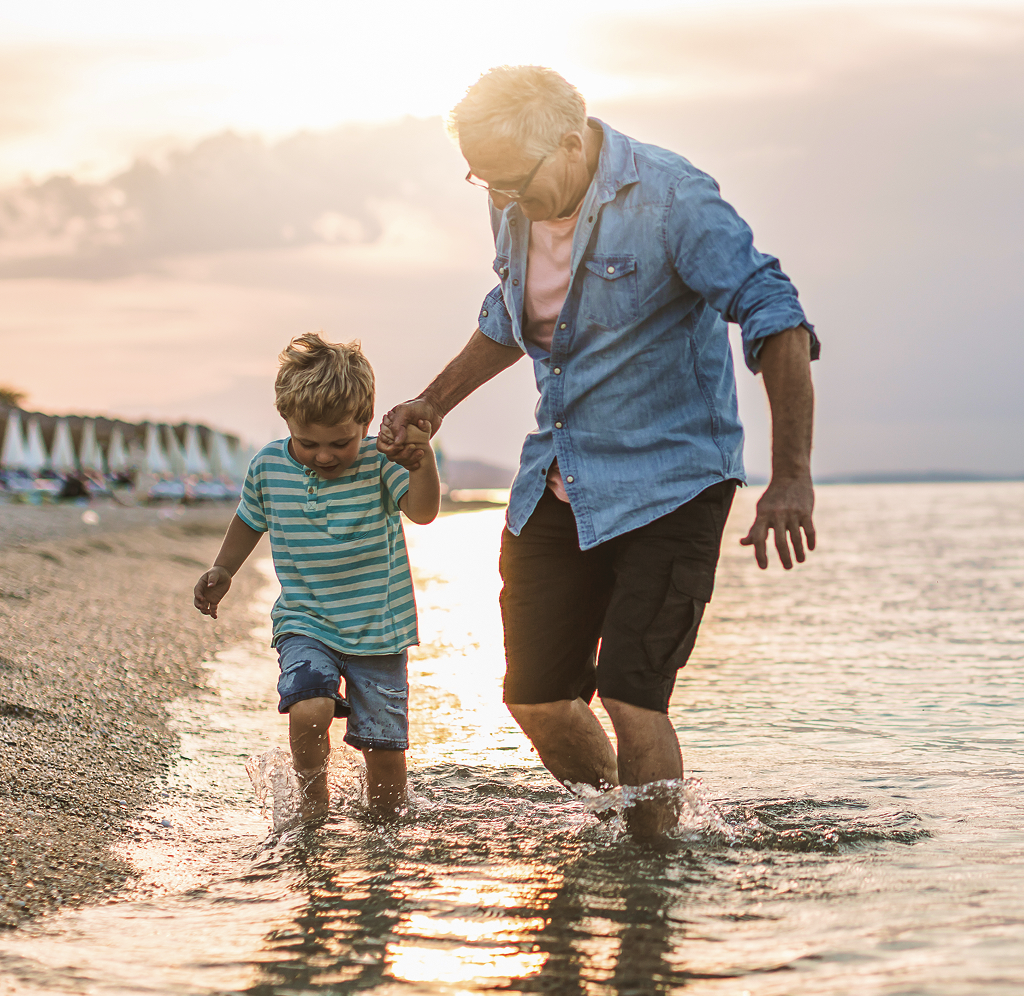 A grandfather running through the water at the beach with his grandchild.