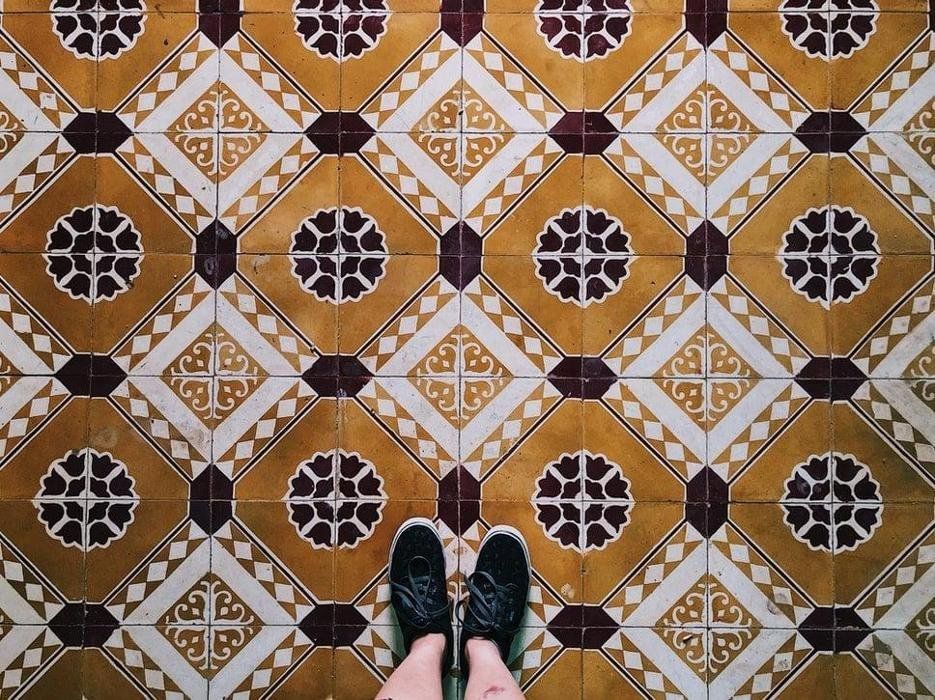 Decorative patterned tile floor with geometric and floral designs in warm tones, viewed from above with person wearing black shoes standing, highlighting vintage style flooring and symmetrical artistic layout.
